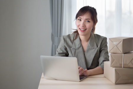 portrait of happy asian woman working with her computer and product parcel box at home; smiling woman business owner works at home or merchandise delivery concept; adult asian chinese woman modelの写真素材