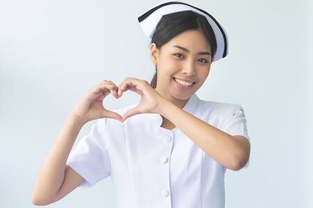 portrait of smiling asian woman nurse with heart hand sign isolated; friendly professional female nurse or medical worker with heart gesture for heart care service or body care or health care conceptの写真素材