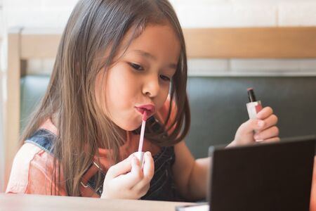 cute little girl playing and applying makeup lipstick to her lips; concept of girl grooming, face care, beauty face, kid playing with cosmetics and lipstick; asian caucasian kid girl modelの写真素材