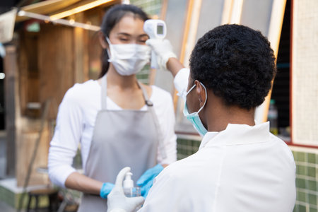 African black woman retail business staff or grocery shop keeper checking fever of Asian coworker by...