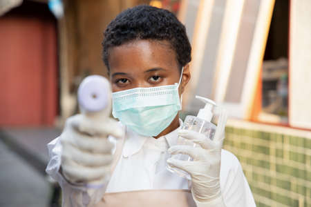 Black African woman employee or shop keeper holding alcohol gel dispenser, hand sanitizer and thermal scanner gun for temperature check; new normal business practice conceptの写真素材