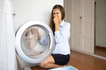 unhappy woman doing laundry howsework, washing dirty stinking cloth with laundry machineの写真素材
