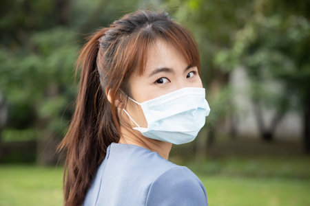 Asian woman wearing face mask looking over shoulder in outdoor park, concept of taking precaution...