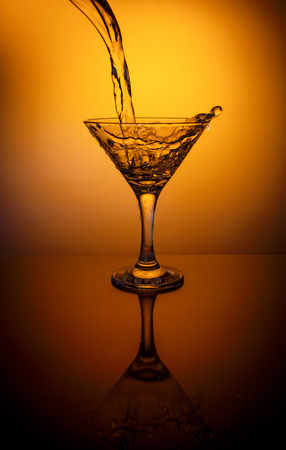 Pouring water from bottle into glass on a thin stalk on orange background, wineglass with reflectionの写真素材