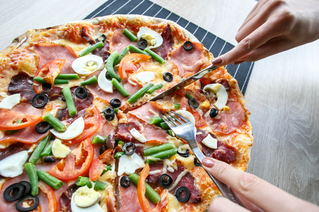 woman hands Slicing fresh pizza with beans, cheese, ham, eggs, pepperoni and vegetables. Cutting pizzaの写真素材