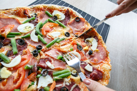 woman hands Slicing fresh pizza with beans, cheese, ham, eggs, pepperoni and vegetables. Cutting pizzaの写真素材