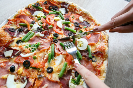 woman hands Slicing fresh pizza with beans, cheese, ham, eggs, pepperoni and vegetables. Cutting pizzaの写真素材