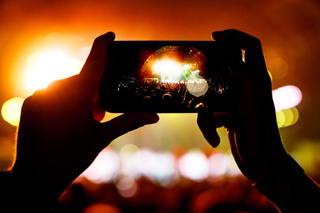 Fan taking photo of concert at festival by smatphone. Arched stage on screenの写真素材