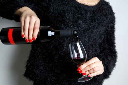Close up of the hands of a young woman pouring red wine into a glass from a bottle, red manicure on the fingersの写真素材