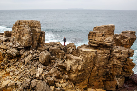 Man stands on a sea cliff, dark stormy sea on background. Young guy travel on the sea and stay on rocky shore under cloudy skyの写真素材