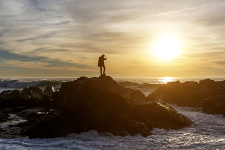 man taking photos of waves on a sea cliff by smartphone, sunsetの写真素材