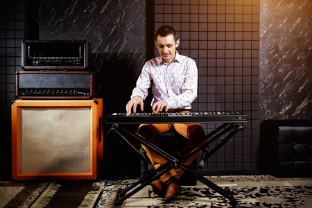 A musician in a light shirt plays a piano synthesizer in a dark recording studio. Amplifiers nearの写真素材