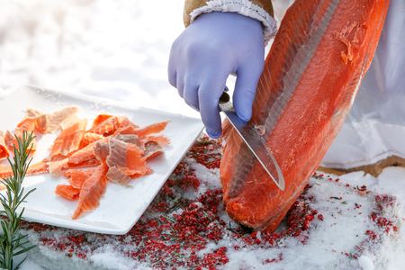 Cutting frozen fish, salmon, on fillets. Red kitchen board, hands in special blue gloves.の写真素材