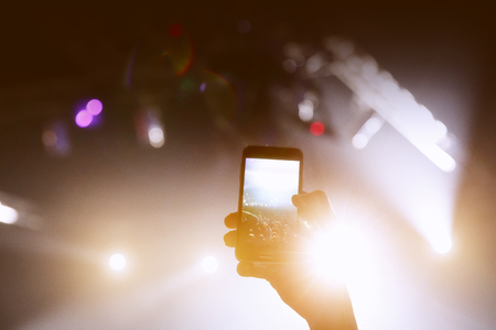 Abstract colorful background of hand holding smartphone to capture image photo and record video in concert music event, black silhouetteの写真素材