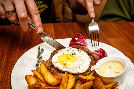 Close-up of hands that cut the beefsteak with scrambled eggs and fried potatoes on the white plate. Side view. Serving for a cafe, a restaurant in the menuの写真素材