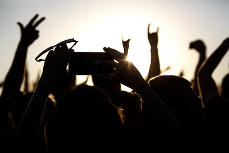 Shooting on a mobile phone during the summer music festival. Black silhouette of raised hands, sunsetの写真素材