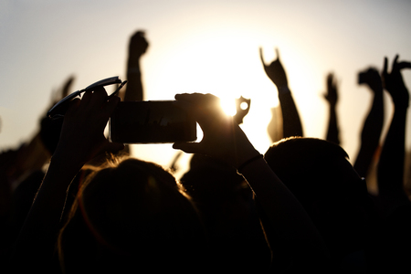 Shooting on a mobile phone during the summer music festival. Black silhouette of raised hands, sunsetの写真素材