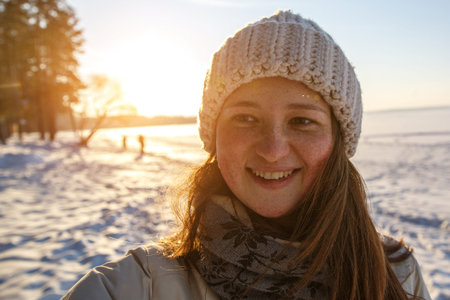 Smiling girl in a warm white knitted hat at winter sunset. Snow background.