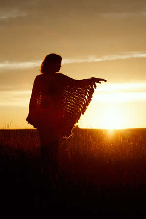 silhouette of happy young woman on sunset, outdoor girl in a plaid poncho in a field with spikeletsの写真素材