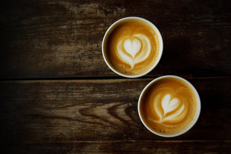 Two disposable cups of coffee with the heart on foam on a wooden tableの写真素材