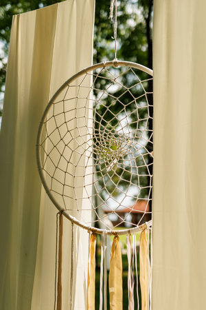 Dreamcatcher, crafted with a white web-like pattern and adorned with white and brown feathers, hanging in front of a window.の写真素材