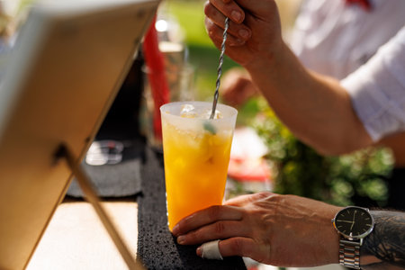 A professional bartender mixes a refreshing orange cocktail with ice and a metal straw in a summer outdoor barの写真素材
