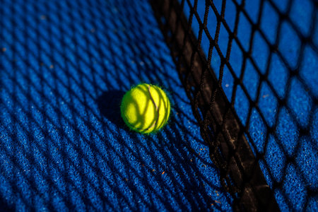 A close-up of a yellow tennis ball near a black net on a blue court surface.の写真素材