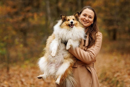 A joyful woman is happily posing with her fluffy collie dog among the vibrant and colorful autumn leavesの写真素材