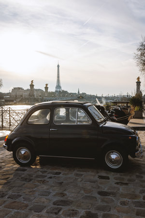 A vintage black car is parked on a cobblestone street in front of the iconic Eiffel Towerの写真素材