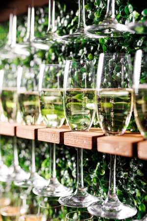 A row of wine glasses are lined up on a shelf. The glasses are all clear and filled with a light