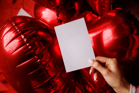 Minimalistic white card held by a hand with red heart-shaped balloons in the background. Suitable...