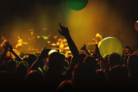 Audience members raise hands during a concert while green and yellow lights glow on stage. The picture captures emotion, enthusiasm, and collective rhythm of live music eventsの写真素材