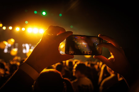 A concertgoer records the performance on a smartphone surrounded by golden stage lighting. The warm colors and silhouettes reflect the energy and emotion of a live event.の写真素材