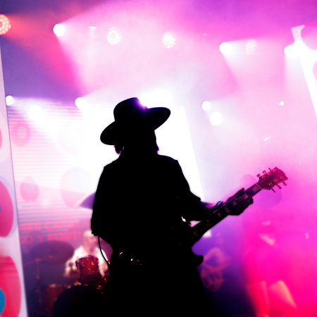 Silhouette of a guitarist wearing a hat performing on stage with bright purple and pink lights.の写真素材