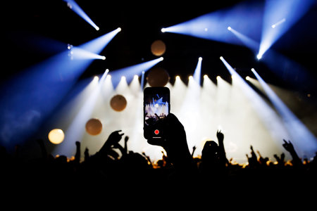 A concert audience records the live performance on a smartphone as bright stage lights and smoke...