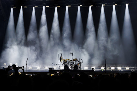 Powerful beams of light illuminate a concert stage with a drum set before the show begins, surrounded by audience silhouettes.の写真素材