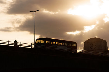 Silhouette of a red bus driving across a London bridge during sunset with cloudy sky and warm light.の写真素材