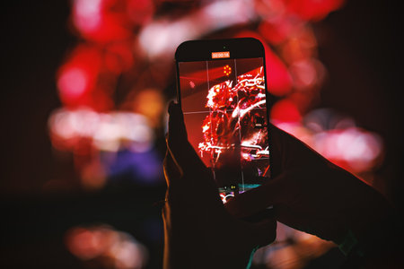 Close-up of hands holding smartphone recording concert performance with red stage lights in the background.の写真素材