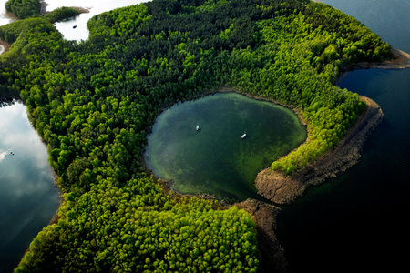 A high-angle drone shot of a green forested peninsula extending into a blue lake with boats on calm water. The image captures the beauty of natural landscapes and untouched wilderness.の写真素材