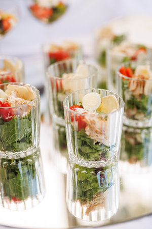 Individual servings of fresh salad with vegetables, egg, and chicken arranged in transparent glass cups.の写真素材