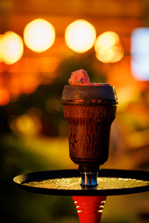 Close-up of a hookah bowl with burning charcoal against a blurred warm background of evening lights.の写真素材