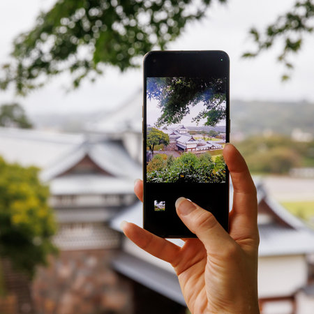 Hand holding a smartphone captures a scenic view of a Japanese castle framed by greenery. The blurred background contrasts with the focused screen.の写真素材