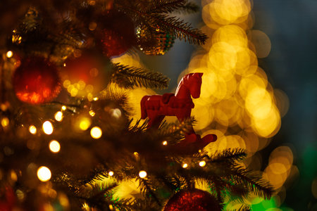 A small, stylized red horse figurine hangs as an ornament on a green Christmas tree branch adorned with string lights, set against a background of brilliant, defocused golden bokeh.の写真素材