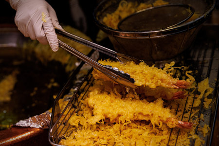 A close-up of a gloved hand using metal tongs to move freshly fried shrimp tempura onto a cooling rack.の写真素材