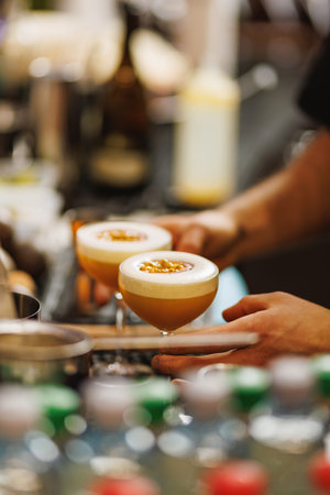 Two passion fruit cocktails in coupe glasses are prepared and served by a bartender. The image captures hands and bar elements in shallow focus.の写真素材