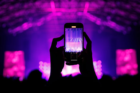 The silhouette of a fan's hands holding a phone vertically to record a stage performance. The entire venue is bathed in vibrant magenta and purple lighting with visible stage screens.の写真素材