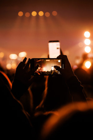 A close-up view of a fan using a mobile phone to record a live musical event. The stage is visible through the phone screen amidst a warm, amber-colored atmosphere.の写真素材