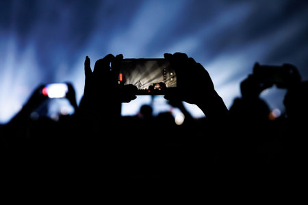Multiple silhouetted fans use their smartphones to capture a live band under dramatic blue stage lights. The venue is dark with bright rays of light cutting through.の写真素材