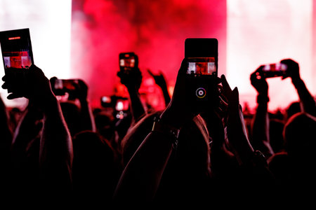 Audience members silhouetted against a vibrant red stage glow with hands holding phones to capture the live event.の写真素材