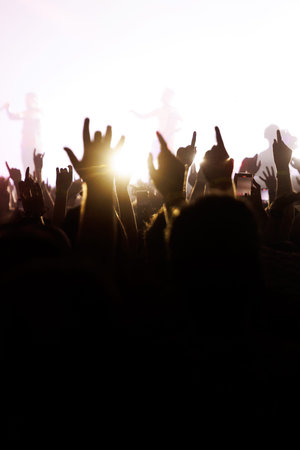 Fans at a rock concert raise their hands in the 'devil horns' gesture against a bright, flared stage light.の写真素材